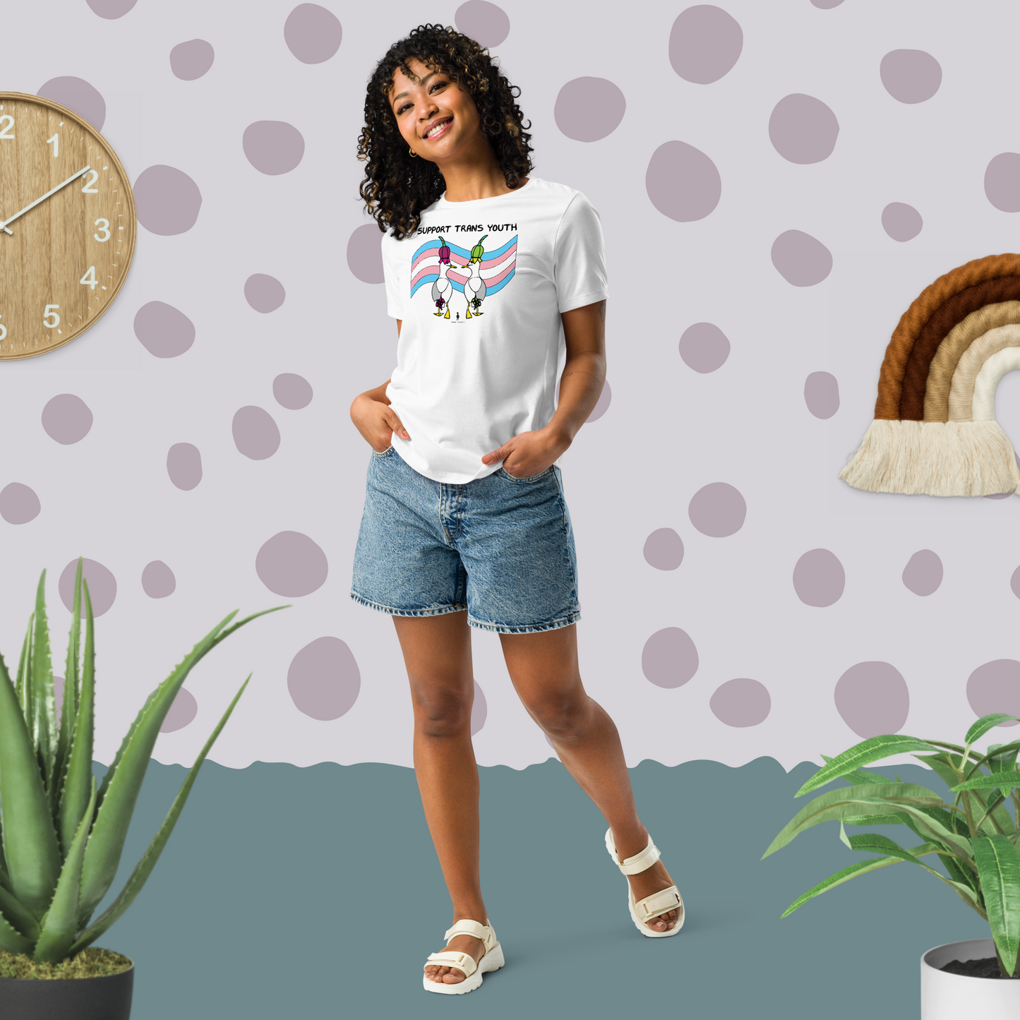 Woman posing casually facing forwards, wearing a fitted white Sassy Seagull Support Trans Youth LGBTQ+ Pride T-Shirt and  stonewash blue jean shorts. She is standing in a pastel coloured boho style room with plants either side and spotty wallpaper. The t-shirt is white with short sleeves. The graphic shows two cartoon style seagulls wearing colourful hats walking away in front of a trans pride flag, underneath the wording Support Trans Youth. It is a soft playful design for everyday wear.