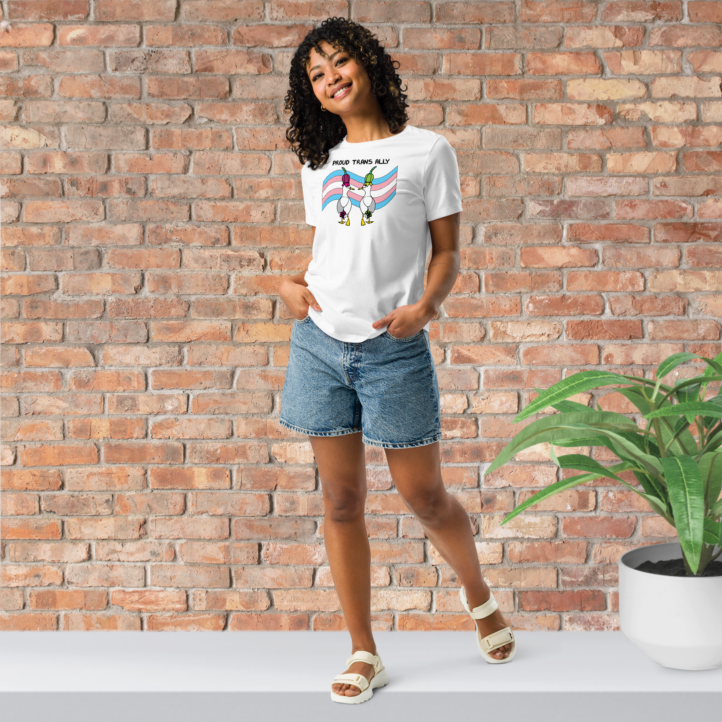Woman posing casually facing forwards, wearing a fitted white Sassy Seagull Proud Trans Ally LGBTQ+ Pride T-Shirt and  stonewash blue jean shorts. She is standing in front of a brick wall with a plant on a white step behind her. The t-shirt is white with short sleeves. The graphic shows two cartoon style seagulls wearing colourful hats walking away in front of an trans pride flag, underneath the wording Proud Trans Ally. It is a soft playful design for everyday wear.