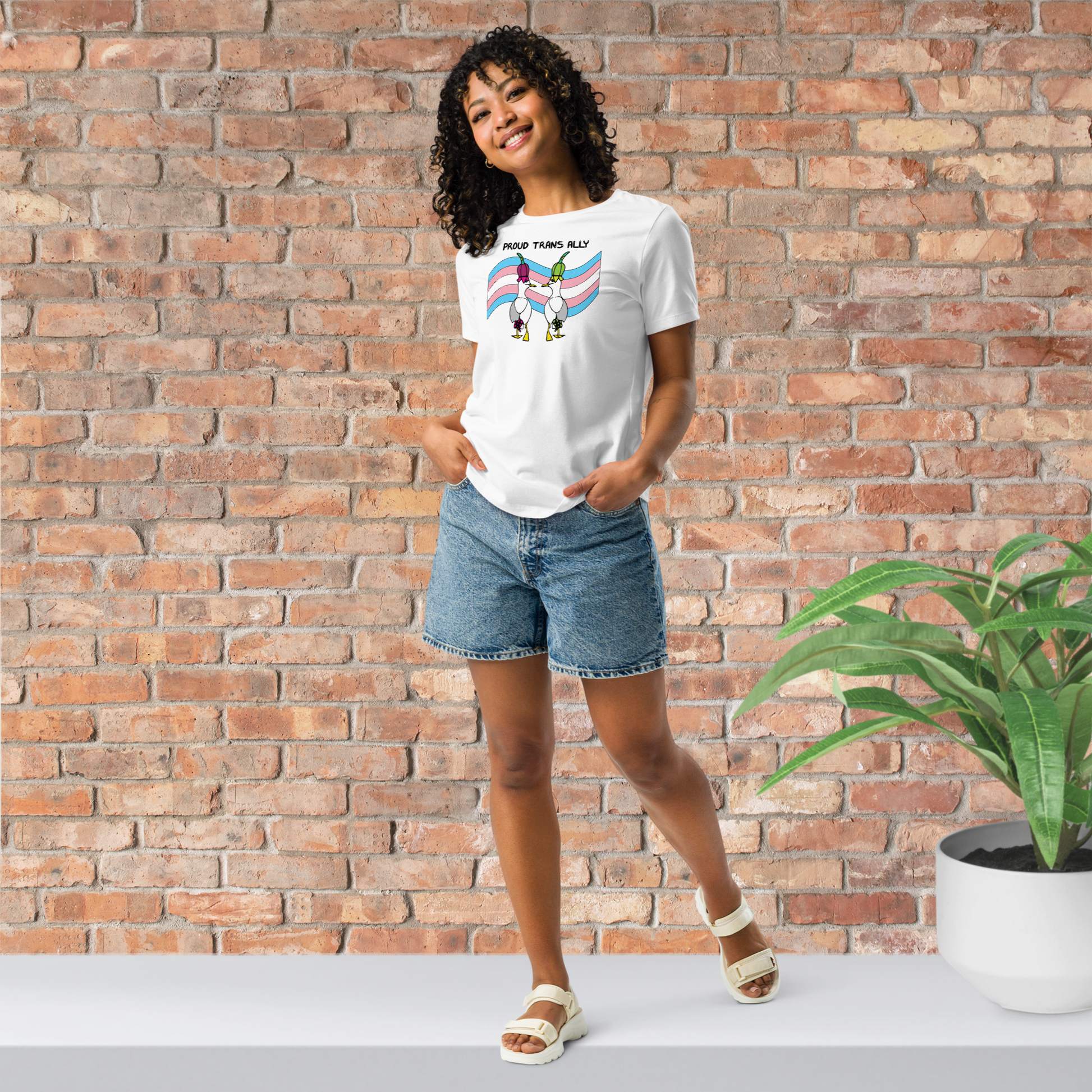 Woman posing casually facing forwards, wearing a fitted white Sassy Seagull Proud Trans Ally LGBTQ+ Pride T-Shirt and  stonewash blue jean shorts. She is standing in front of a brick wall with a plant on a white step behind her. The t-shirt is white with short sleeves. The graphic shows two cartoon style seagulls wearing colourful hats walking away in front of an trans pride flag, underneath the wording Proud Trans Ally. It is a soft playful design for everyday wear.