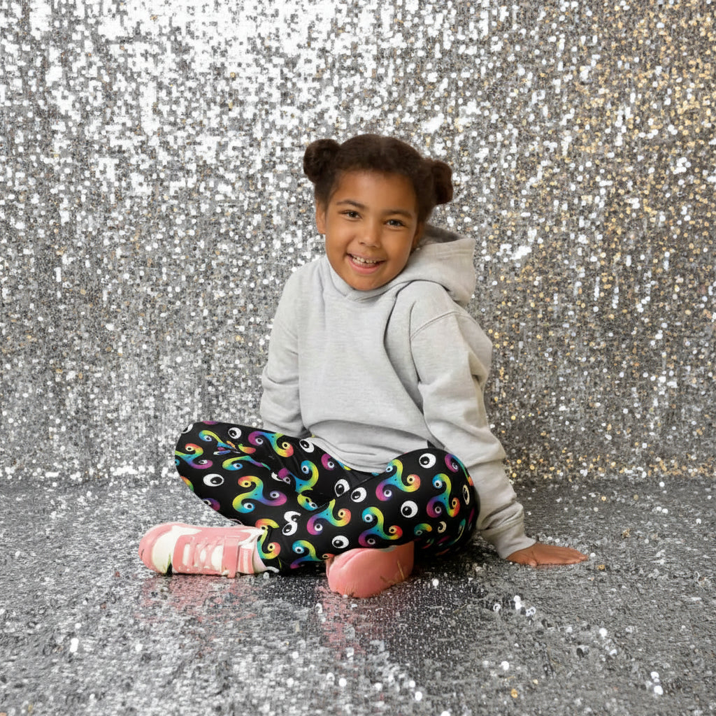 Young girl sitting cross legged in a country garden full of flowers, smiling at the camera, wearing a grey hoodie, trippy triskelion leggings and pink trainers
