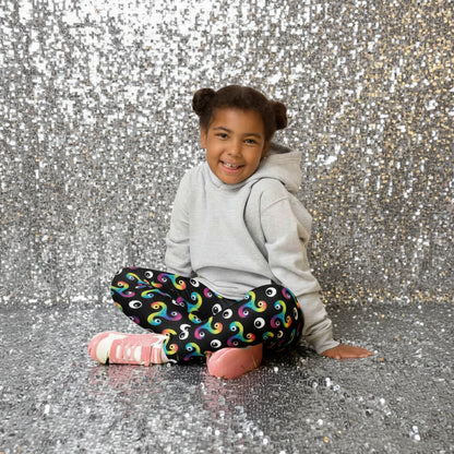 Young girl sitting cross legged in a country garden full of flowers, smiling at the camera, wearing a grey hoodie, trippy triskelion leggings and pink trainers