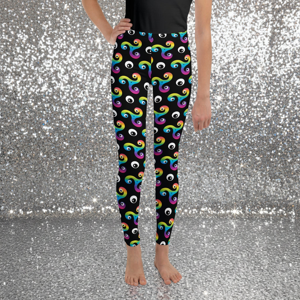 Teen standing facing forward wearing a black t-shirt and trippy triskelion leggings on a sandy beach with palm trees