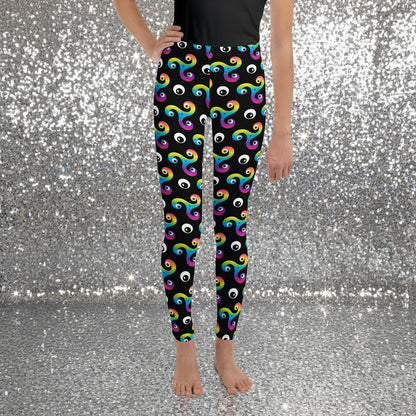 Teen standing facing forward wearing a black t-shirt and trippy triskelion leggings on a sandy beach with palm trees