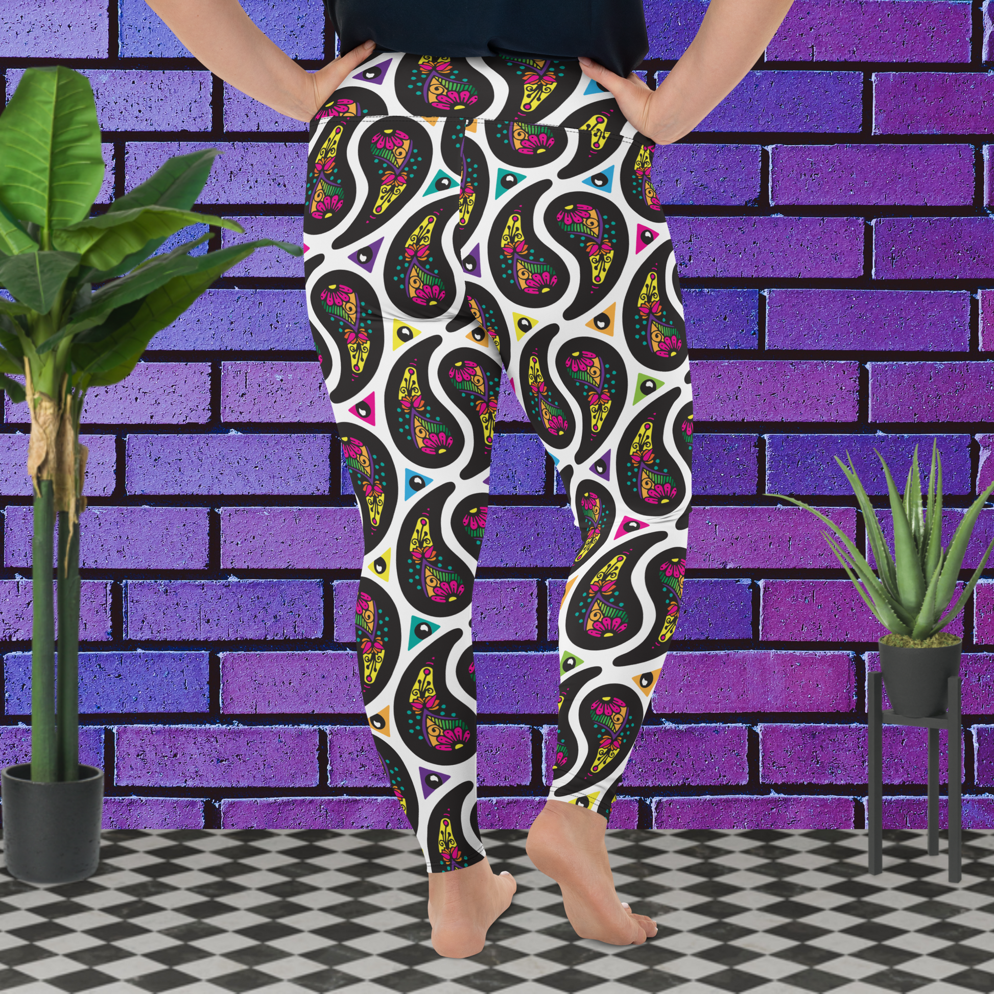 A curvy woman from the waist down wearing plus-size white Paisley Party high-waist funky festival yoga leggings standing facing to the back with her hands on her hips. The woman is standing on a black and white tiled floor in front of a vivid purple brick wall with 2 leafy green plants on the floor, one on either side.The Paisley Party pattern is a repeating paisley pattern. The paisley shapes are filled with decorative detail in a variety of bright colours against a white background.