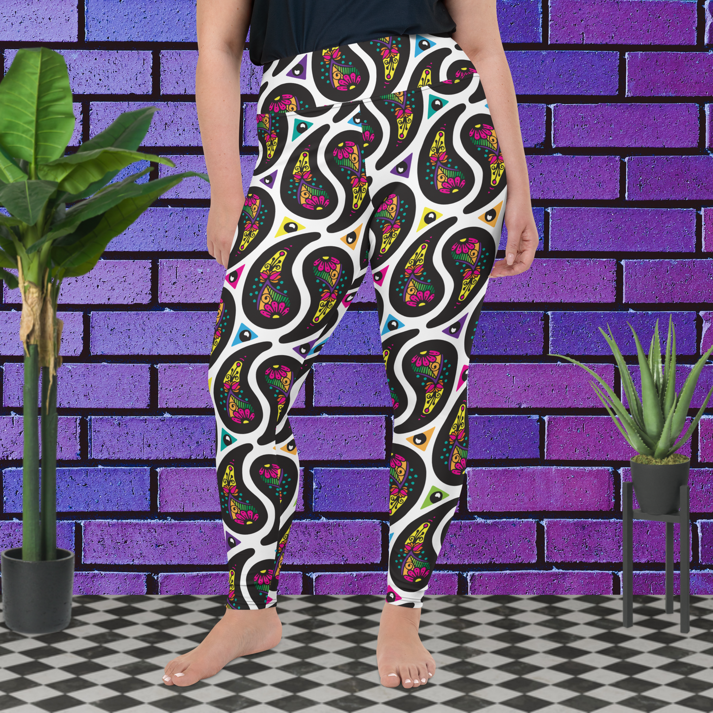 A curvy woman from the waist down wearing plus-size white Paisley Party high-waist funky festival yoga leggings standing facing forwards with her hands by her sides. The woman is standing on a black and white tiled floor in front of a vivid purple brick wall with 2 leafy green plants on the floor, one on either side.The Paisley Party pattern is a repeating paisley pattern. The paisley shapes are filled with decorative detail in a variety of bright colours against a white background.
