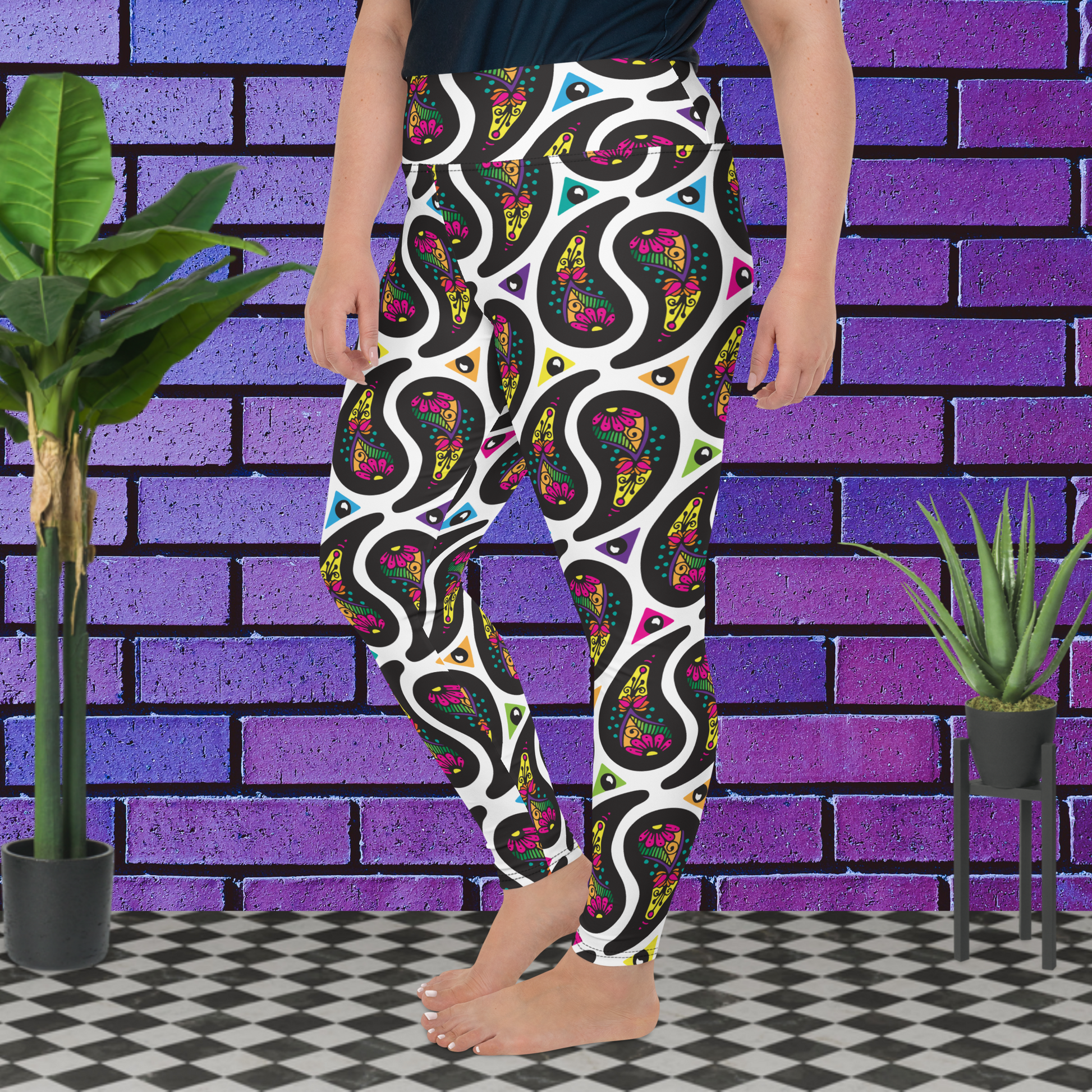 A curvy woman from the waist down wearing plus-size white Paisley Party high-waist funky festival yoga leggings standing facing slightly to the left with her hands by her sides. The woman is standing on a black and white tiled floor in front of a vivid purple brick wall with 2 leafy green plants on the floor, one on either side.The Paisley Party pattern is a repeating paisley pattern. The paisley shapes are filled with decorative detail in a variety of bright colours against a white background.