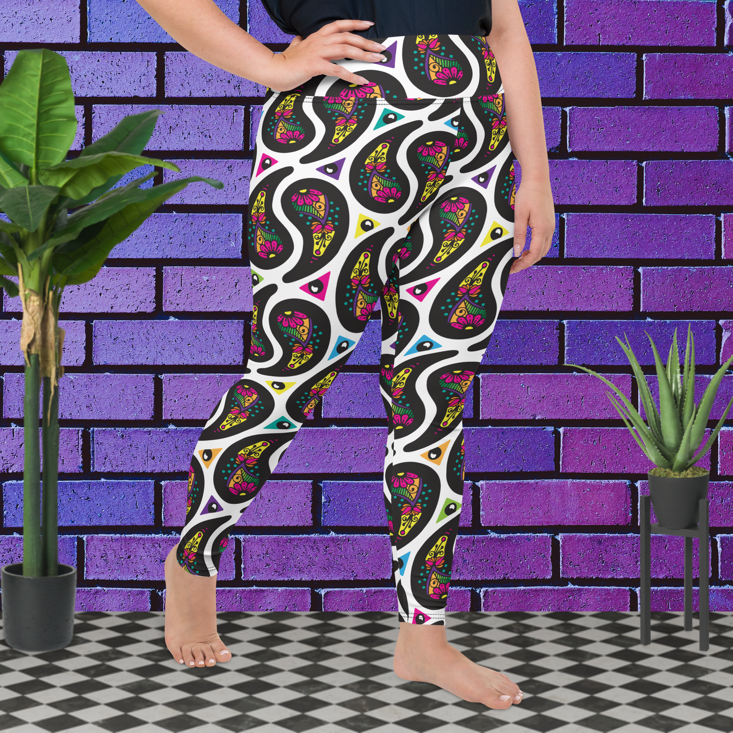 A curvy woman from the waist down wearing plus-size white Paisley Party high-waist funky festival yoga leggings standing facing slightly to the right with her  right hand on her hip. The woman is standing on a black and white tiled floor in front of a vivid purple brick wall with 2 leafy green plants on the floor, one on either side.The Paisley Party pattern is a repeating paisley pattern. The paisley shapes are filled with decorative detail in a variety of bright colours against a white background.