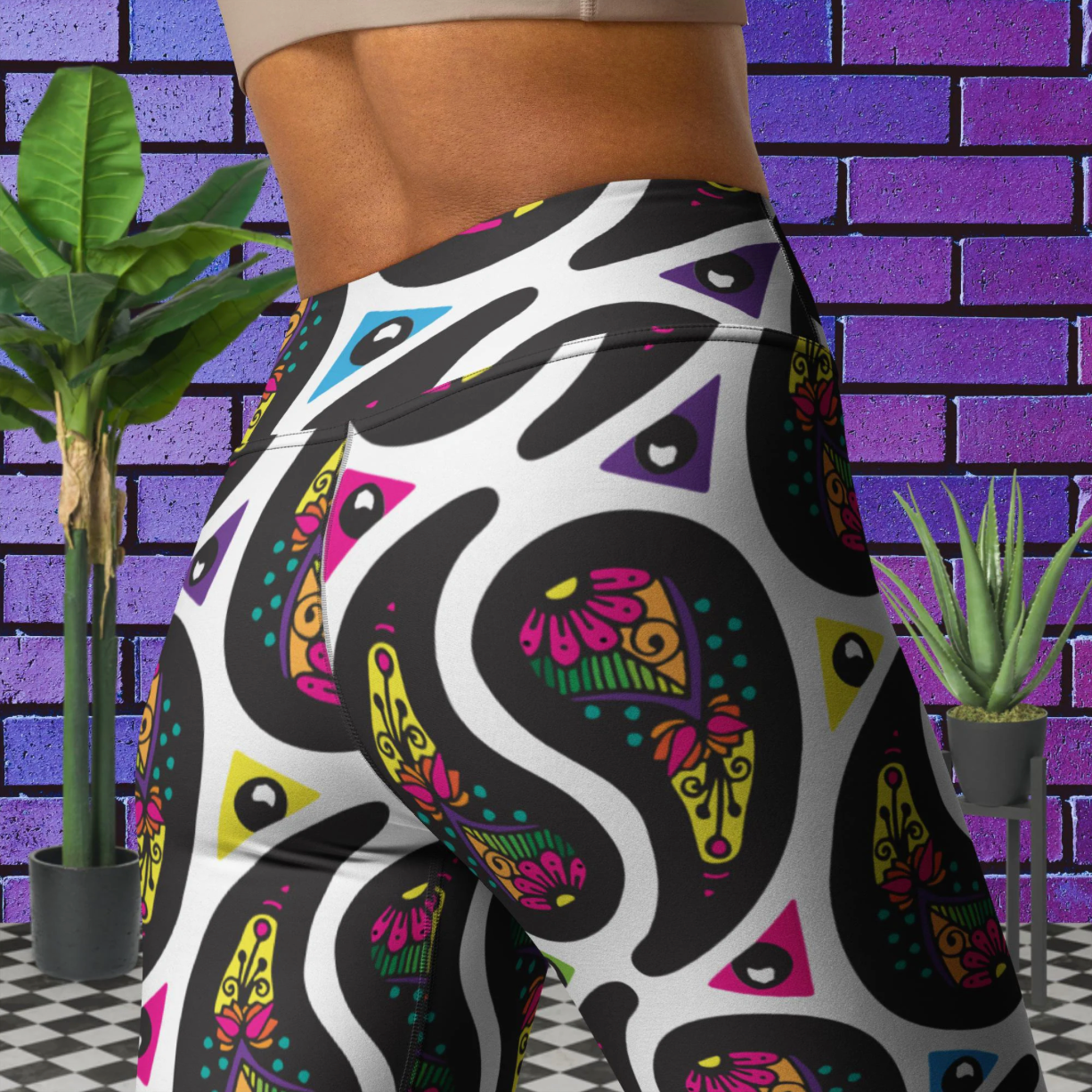 Close up of a woman from the chest down to her thighs wearing white Paisley Party high-waist funky festival yoga leggings standing facing backwards and slightly to the right. The woman is standing on a black and white tiled floor in front of a vivid purple brick wall with 2 leafy green plants on the floor, one on either side.The Paisley Party pattern is a repeating paisley pattern. The paisley shapes are filled with decorative detail in a variety of bright colours against a white background.