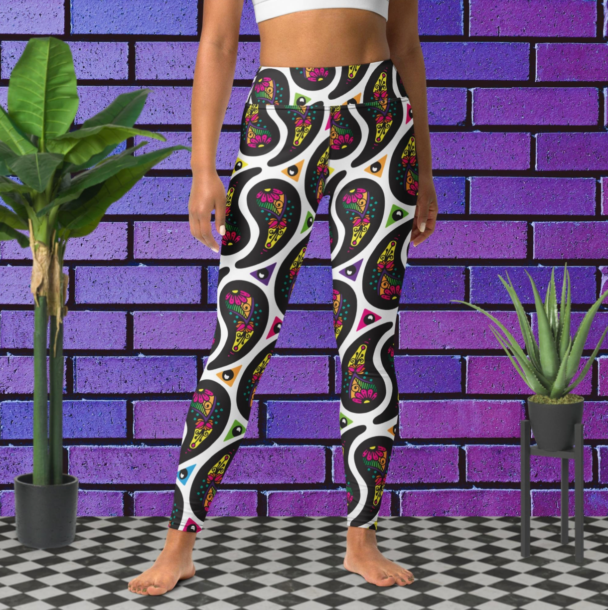 Woman from the waist down wearing white Paisley Party high-waist funky festival yoga leggings standing facing forwards with her hands by her sides. The woman is standing on a black and white tiled floor in front of a vivid purple brick wall with 2 leafy green plants on the floor, one on either side.The Paisley Party pattern is a repeating paisley pattern. The paisley shapes are filled with decorative detail in a variety of bright colours against a white background.