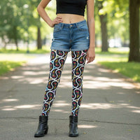 A young person poses facing forwards with one hand on their hip. They are standing in an interior space which looks mediterranean-esque. There are leafy plants. They are wearing white paisley party funky festival leggings, and a black t-shirt. The paisley party pattern  is a colourful repeating large paisley pattern, made up of vibrant colours including pink, orange, yellow, green, blue and purple. In between the paisley print are colourful triangles in the same colours. The pattern  background is white.