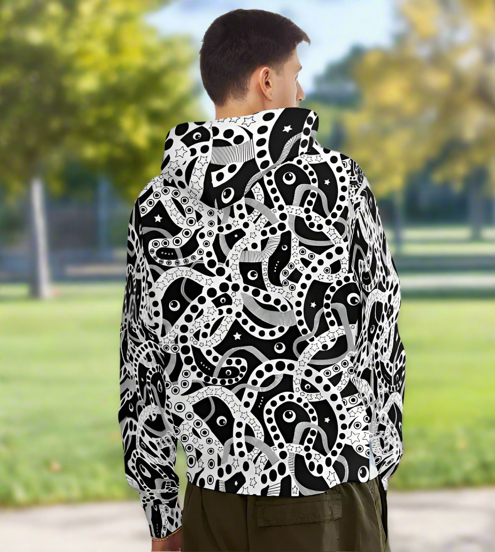 Man from the chin down to the waist, posing standing facing away wearing a 100% cotton bold black and white Who's Afraid Of The Dark all-over print hoodie. He is standing in a park with trees in the background. The hoodie has an abstract design made up of interwoven shapes detailed with spots, stars and lines. It is a unique and bold design for everyday wear.