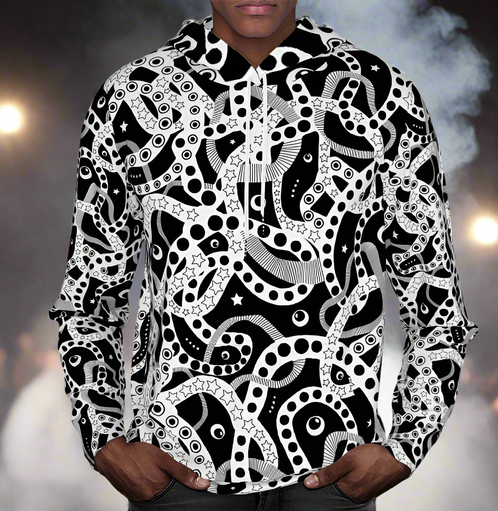 Man from the chin down to the waist, posing standing facing forwards wearing a 100% cotton bold black and white Who's Afraid Of The Dark all-over print hoodie. He is standing against a stage background with lights and smoke effects. The hoodie has an abstract design made up of interwoven shapes detailed with spots, stars and lines. It is a unique and bold design for everyday wear.