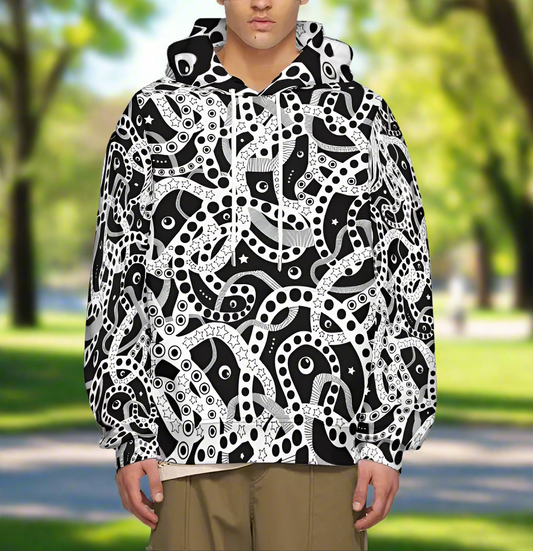 Man from the chin down to the waist, posing standing facing forwards wearing a 100% cotton bold black and white Who's Afraid Of The Dark all-over print hoodie. He is standing in a park surrounded by trees. The hoodie has an abstract design made up of interwoven shapes detailed with spots, stars and lines. It is a unique and bold design for everyday wear.