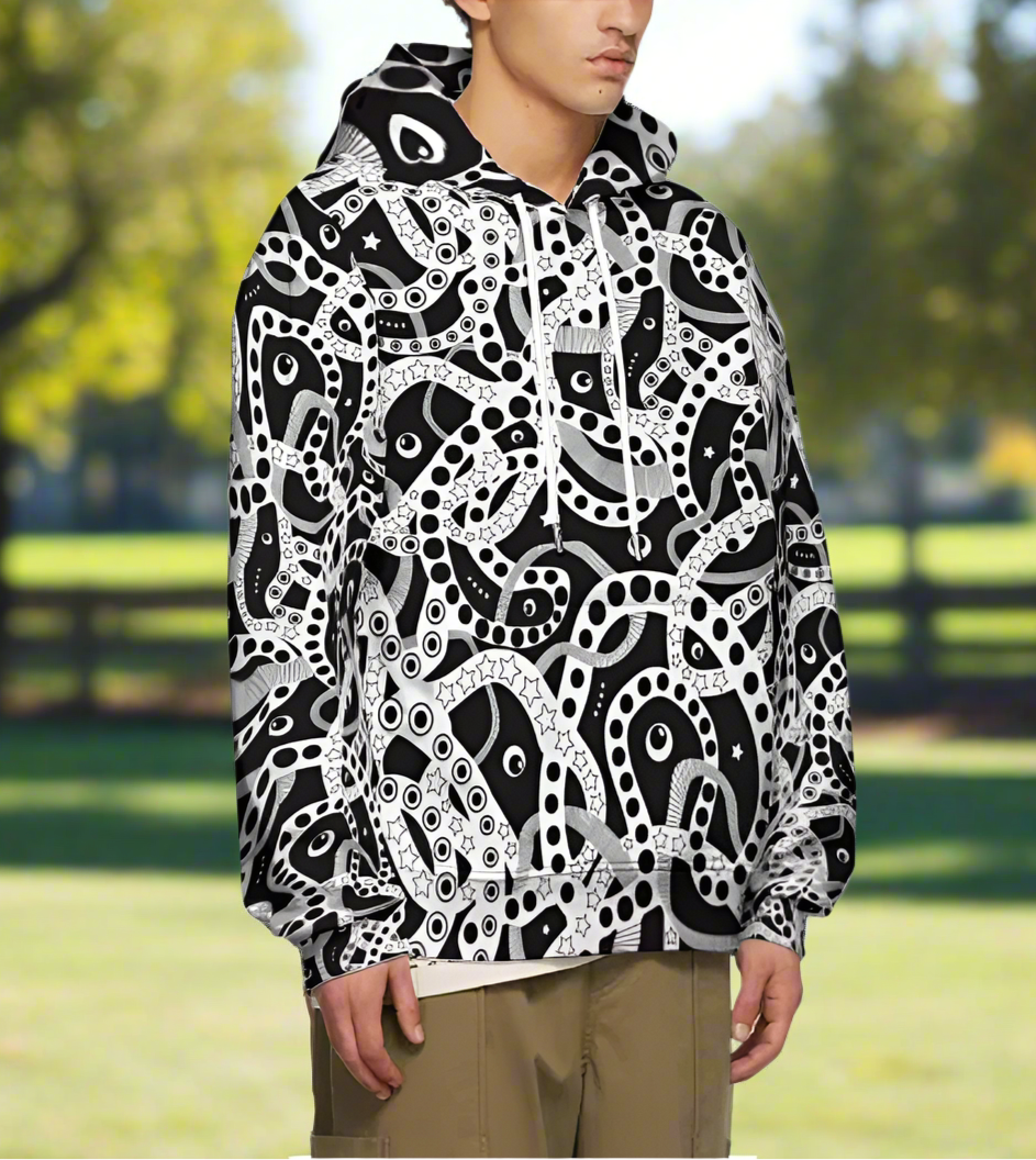 Man from the chin down to the waist, posing standing facing to the right wearing a 100% cotton bold black and white Who's Afraid Of The Dark all-over print hoodie. He is standing in a park with trees in the background. The hoodie has an abstract design made up of interwoven shapes detailed with spots, stars and lines. It is a unique and bold design for everyday wear.