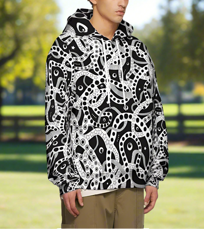 Man from the chin down to the waist, posing standing facing to the right wearing a 100% cotton bold black and white Who's Afraid Of The Dark all-over print hoodie. He is standing in a park with trees in the background. The hoodie has an abstract design made up of interwoven shapes detailed with spots, stars and lines. It is a unique and bold design for everyday wear.