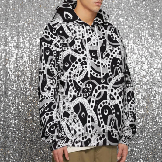 Man from the chin down to the waist, posing standing facing to the right wearing a 100% cotton bold black and white Who's Afraid Of The Dark all-over print hoodie. He is standing in a park with trees in the background. The hoodie has an abstract design made up of interwoven shapes detailed with spots, stars and lines. It is a unique and bold design for everyday wear.