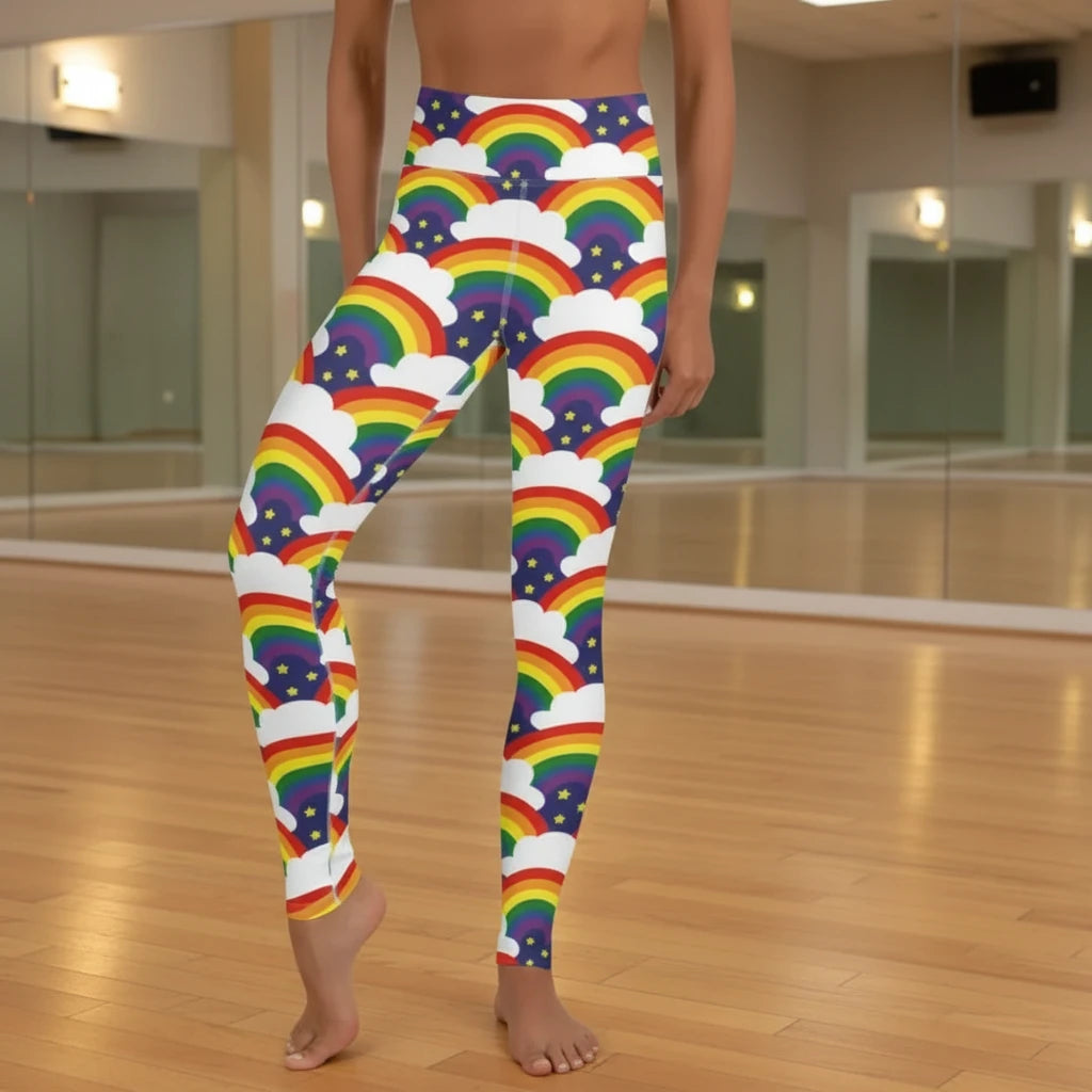 High waist Pride Rainbow Sparkle festival yoga leggings front view. The woman is standing in a rustic boho setting with plants, fairy lights and bunting in the background.  The Rainbow Sparkle design is a bold rainbow, clouds and stars pattern in the LGBTQ+ pride palette. Designed in Wales, UK | Sassy Seagull. 