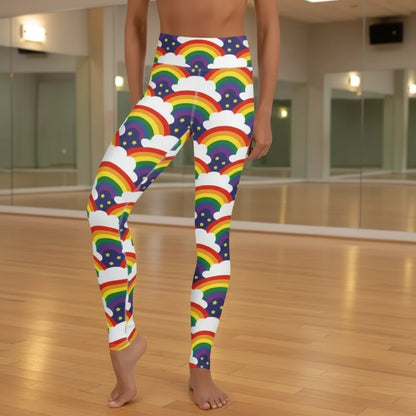 High waist Pride Rainbow Sparkle festival yoga leggings front view. The woman is standing in a rustic boho setting with plants, fairy lights and bunting in the background.  The Rainbow Sparkle design is a bold rainbow, clouds and stars pattern in the LGBTQ+ pride palette. Designed in Wales, UK | Sassy Seagull. 