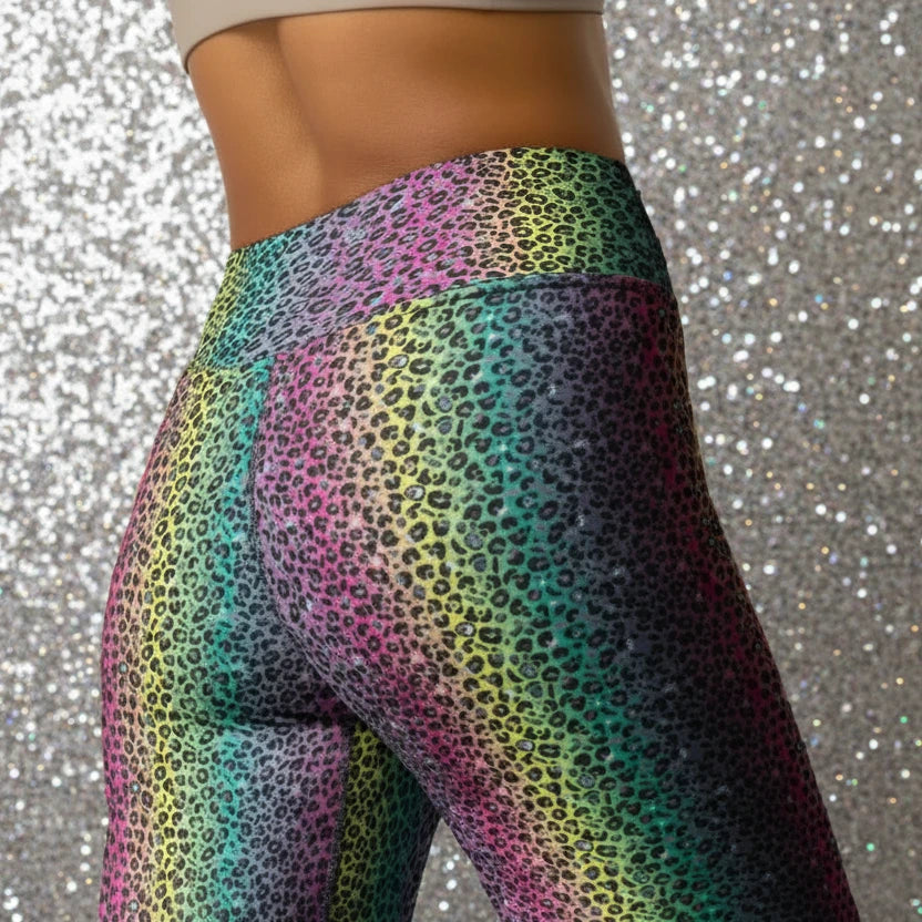 High waist Rainbow Leopard Glamour festival yoga leggings close up back view. The woman is standing in a rustic boho setting with plants, fairy lights and bunting in the background.  The rainbow leopard glamour design is a bold animal printed pattern in black, grey and white against a blended rainbow background. Designed in Wales, UK | Sassy Seagull. 