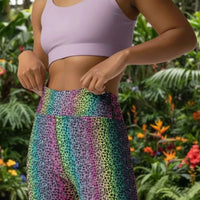 High waist Rainbow Leopard Glamour festival yoga leggings close up front view. The woman is standing in a rustic boho setting with plants, fairy lights and bunting in the background.  The rainbow leopard glamour design is a bold animal printed pattern in black, grey and white against a blended rainbow background. Designed in Wales, UK | Sassy Seagull. 