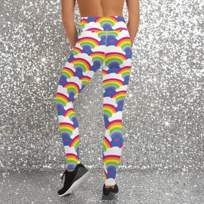 High waist Rainbow Love festival yoga leggings back view. The woman is standing in a rustic boho setting with plants, fairy lights and bunting in the background.  The Rainbow Love design is a bold design featuring rainbows, white clouds and pink hearts. Designed in Wales, UK | Sassy Seagull. 