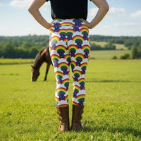 High waist Plus Size Pride Rainbow Sparkle festival yoga leggings back view. The woman is standing in a rustic boho setting with plants, fairy lights and bunting in the background.  The Rainbow Sparkle design is a bold rainbow, clouds and stars pattern in the LGBTQ+ pride palette. Designed in Wales, UK | Sassy Seagull. 