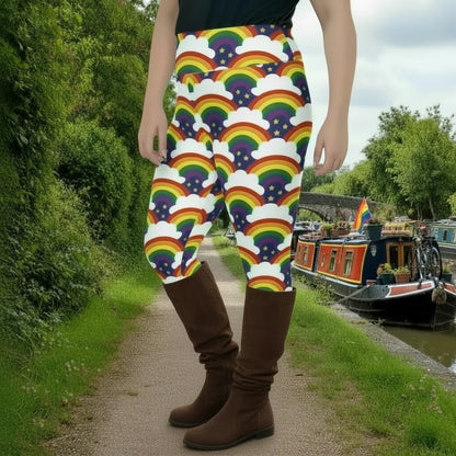 High waist Plus Size Pride Rainbow Sparkle festival yoga leggings left side view. The woman is standing in a rustic boho setting with plants, fairy lights and bunting in the background.  The Rainbow Sparkle design is a bold rainbow, clouds and stars pattern in the LGBTQ+ pride palette. Designed in Wales, UK | Sassy Seagull. 