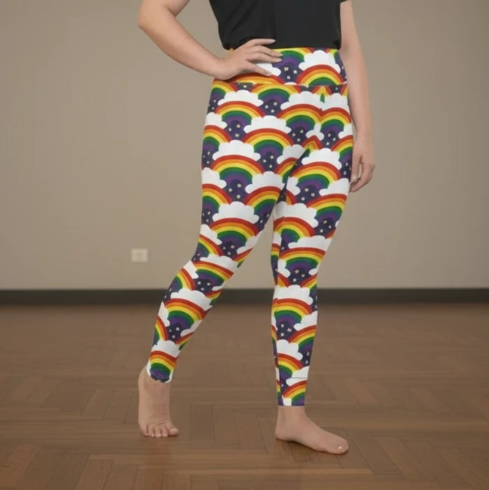 High waist Plus Size Pride Rainbow Sparkle festival yoga leggings right side view. The woman is standing in a rustic boho setting with plants, fairy lights and bunting in the background.  The Rainbow Sparkle design is a bold rainbow, clouds and stars pattern in the LGBTQ+ pride palette. Designed in Wales, UK | Sassy Seagull. 