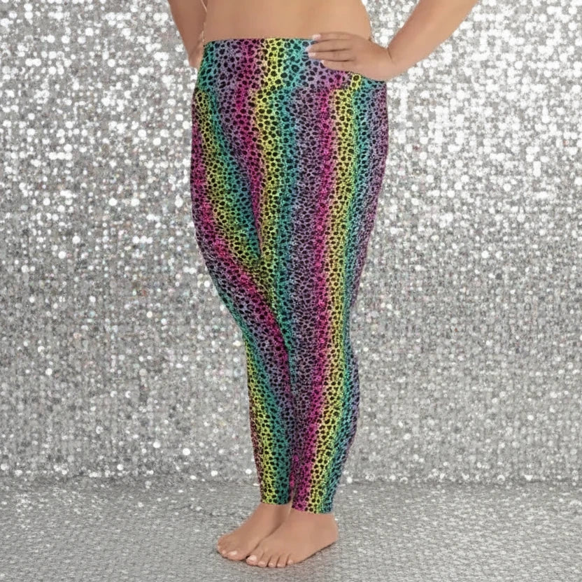High waist Plus Size Rainbow Leopard Glamour festival yoga leggings side view. The woman is standing in a rustic boho setting with plants, fairy lights and bunting in the background.  The rainbow leopard glamour design is a bold animal printed pattern in black, grey and white against a blended rainbow background. Designed in Wales, UK | Sassy Seagull. 