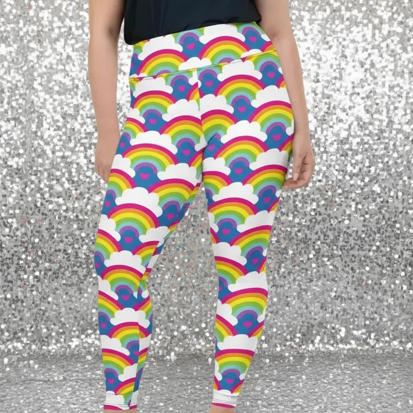 Plus-size high waist Rainbow Love festival yoga leggings front view. The woman is standing in a rustic boho setting with plants, fairy lights and bunting in the background.  The Rainbow Love design is a bold design featuring rainbows, white clouds and pink hearts. Designed in Wales, UK | Sassy Seagull. 