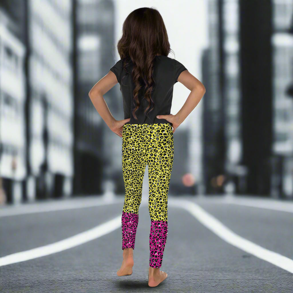 A young girl wearing kids size yellow and pink leopard glamour funky festival leggings and a black t-shirt stands facing away. She is standing in front of a black and white city street scape.  This design features an animal print pattern in black, grey and white, printed at a medium scale. The top 3/4 background is bright yellow with the bottom 1/4 bright pink
