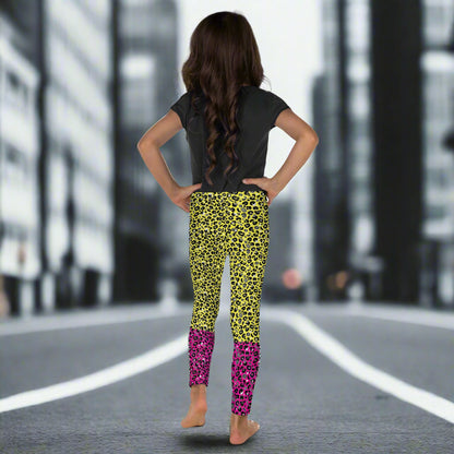 A young girl wearing kids size yellow and pink leopard glamour funky festival leggings and a black t-shirt stands facing away. She is standing in front of a black and white city street scape.  This design features an animal print pattern in black, grey and white, printed at a medium scale. The top 3/4 background is bright yellow with the bottom 1/4 bright pink