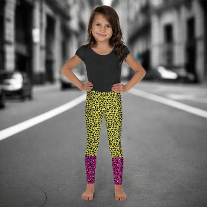 A young girl wearing kids size yellow and pink leopard glamour funky festival leggings and a black t-shirt stands facing forwards. She is smiling and standing in front of a black and white city street scape.  This design features an animal print pattern in black, grey and white, printed at a medium scale. The top 3/4 background is bright yellow with the bottom 1/4 bright pink