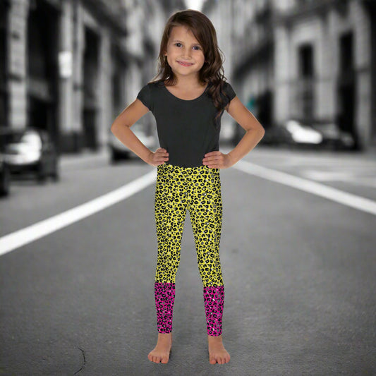 A young girl wearing kids size yellow and pink leopard glamour funky festival leggings and a black t-shirt stands facing forwards. She is smiling and standing in front of a black and white city street scape.  This design features an animal print pattern in black, grey and white, printed at a medium scale. The top 3/4 background is bright yellow with the bottom 1/4 bright pink