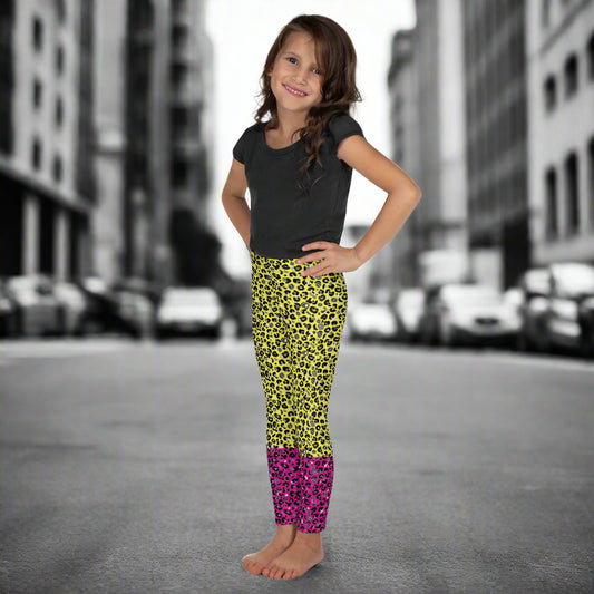 A young girl wearing kids size yellow and pink leopard glamour funky festival leggings and a black t-shirt stands facing to the left. She is smiling and standing in front of a black and white city street scape.  This design features an animal print pattern in black, grey and white, printed at a medium scale. The top 3/4 background is bright yellow with the bottom 1/4 bright pink