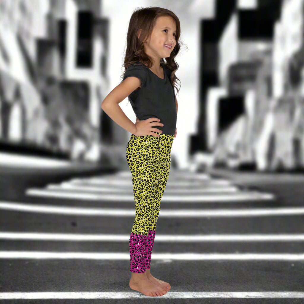 A young girl wearing kids size yellow and pink leopard glamour funky festival leggings and a black t-shirt stands facing to the right. She is smiling and standing in front of a black and white city street scape.  This design features an animal print pattern in black, grey and white, printed at a medium scale. The top 3/4 background is bright yellow with the bottom 1/4 bright pink