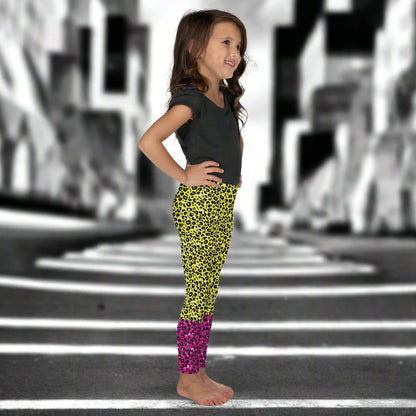 A young girl wearing kids size yellow and pink leopard glamour funky festival leggings and a black t-shirt stands facing to the right. She is smiling and standing in front of a black and white city street scape.  This design features an animal print pattern in black, grey and white, printed at a medium scale. The top 3/4 background is bright yellow with the bottom 1/4 bright pink