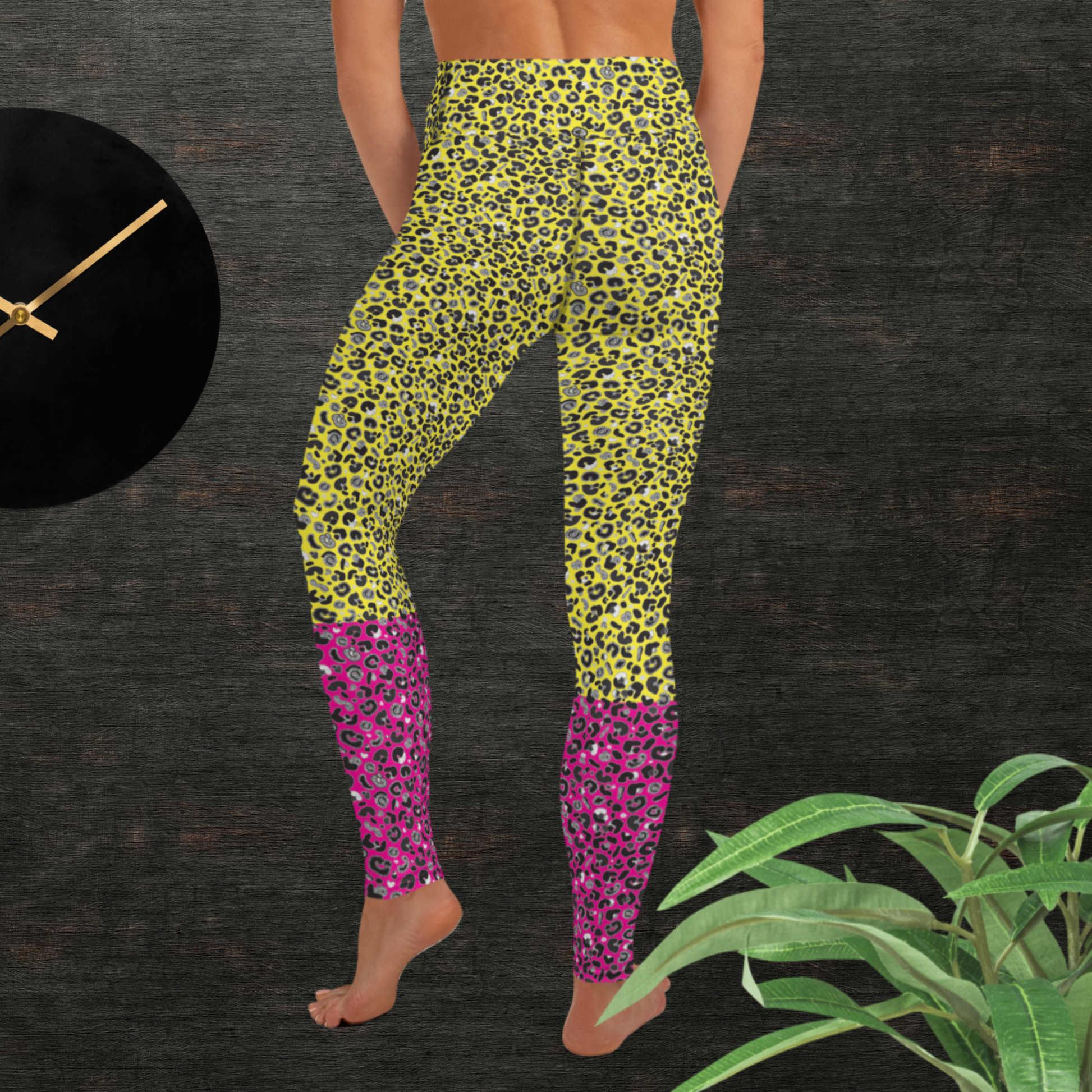 Woman from the waist down wearing high-waist Yellow and Pink Leopard Glamour funky festival yoga leggings standing facing away with her hands in front of her. The woman is standing in front of a textured grey background with a black and gold wall clock to the left and a partially visible leafy plant on the floor in the bottom right.  This design features an animal print pattern in black, grey and white, printed at a medium scale. The top 3/4 background is bright yellow with the bottom 1/4 a hot pink colour
