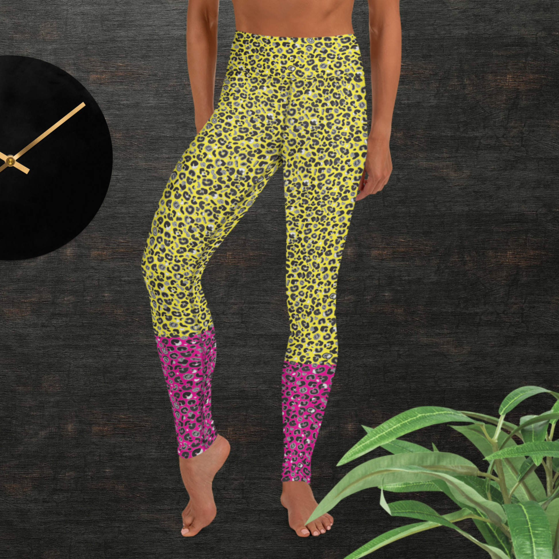 Woman from the waist down wearing high-waist Yellow and Pink Leopard Glamour funky festival yoga leggings standing facing forwards with her hands by her sides. The woman is standing in front of a textured grey background with a black and gold wall clock to the left and a partially visible leafy plant on the floor in the bottom right.  This design features an animal print pattern in black, grey and white, printed at a medium scale. The top 3/4 background is bright yellow with the bottom 1/4 a hot pink colour