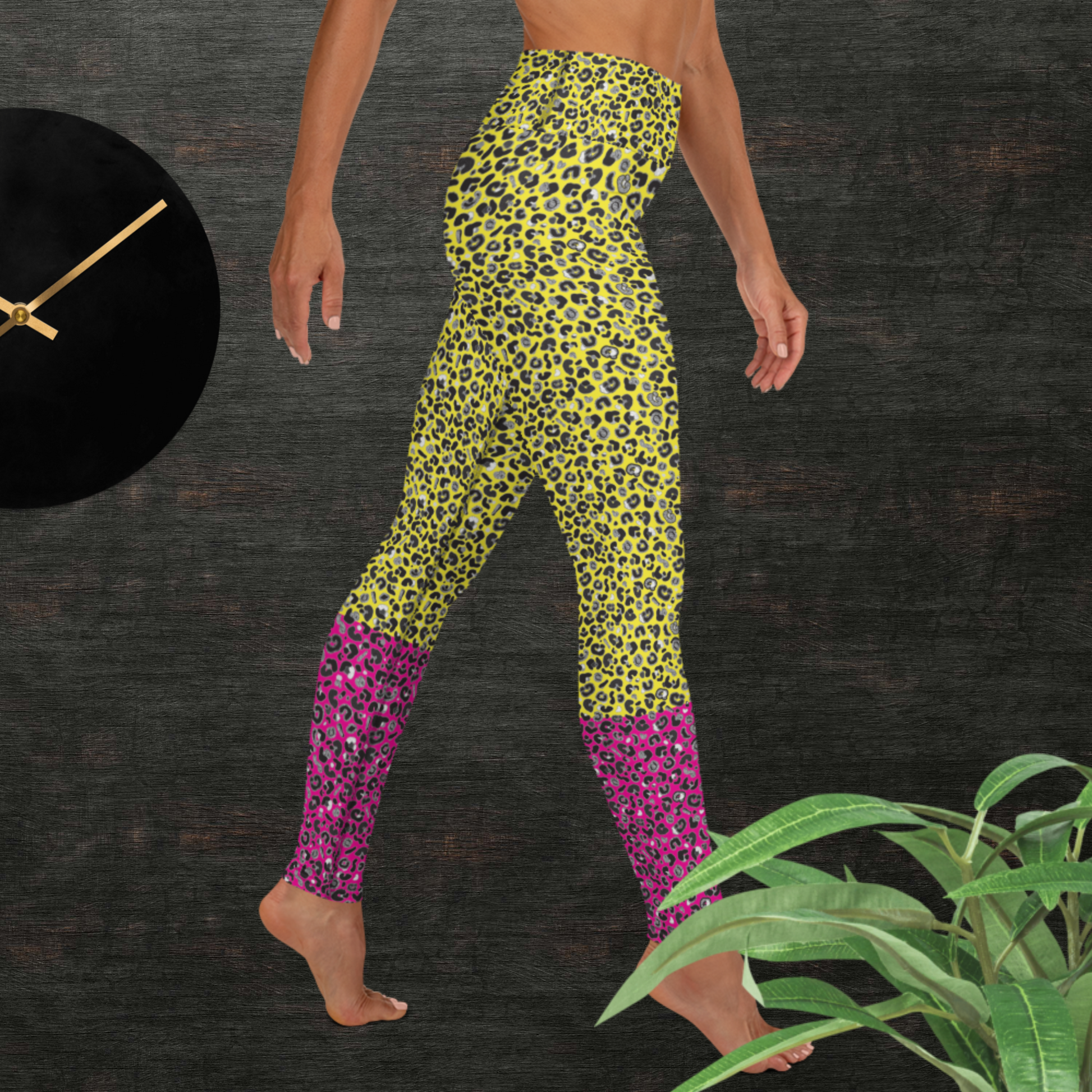 Woman from the waist down wearing high-waist Yellow and Pink Leopard Glamour funky festival yoga leggings standing facing right with her hands by her sides. The woman is standing in front of a textured grey background with a black and gold wall clock to the left and a partially visible leafy plant on the floor in the bottom right.  This design features an animal print pattern in black, grey and white, printed at a medium scale. The top 3/4 background is bright yellow with the bottom 1/4 a hot pink colour