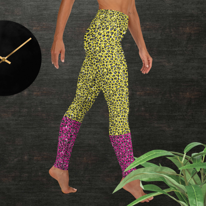 Woman from the waist down wearing high-waist Yellow and Pink Leopard Glamour funky festival yoga leggings standing facing right with her hands by her sides. The woman is standing in front of a textured grey background with a black and gold wall clock to the left and a partially visible leafy plant on the floor in the bottom right.  This design features an animal print pattern in black, grey and white, printed at a medium scale. The top 3/4 background is bright yellow with the bottom 1/4 a hot pink colour