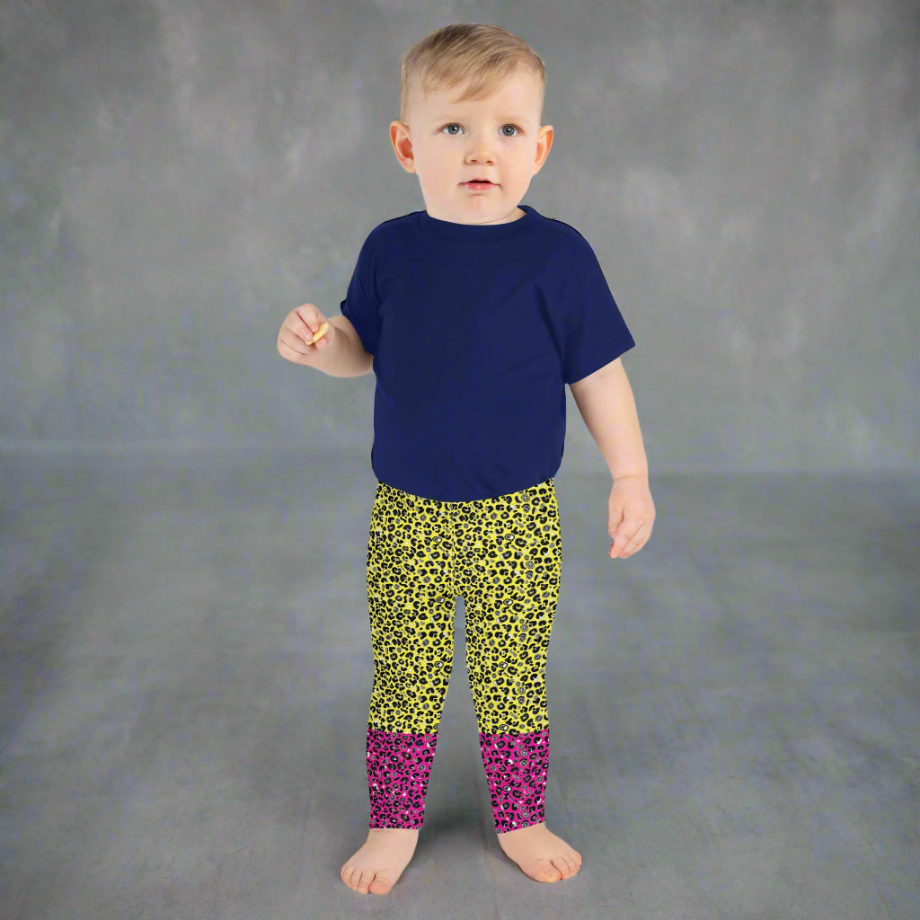 A toddler wearing kids size yellow and pink leopard glamour funky festival leggings and a navy t-shirt stands facing forwards. He is standing facing forwards in front of a textured grey background  This design features an animal print pattern in black, grey and white, printed at a medium scale. The top 3/4 background is bright yellow with the bottom 1/4 bright pink