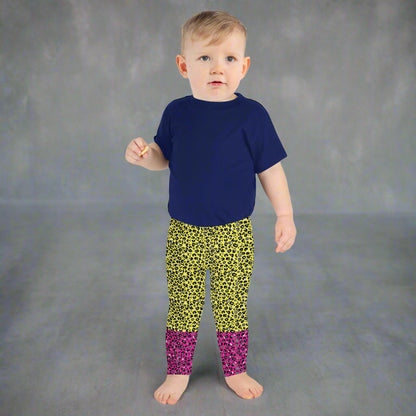 A toddler wearing kids size yellow and pink leopard glamour funky festival leggings and a navy t-shirt stands facing forwards. He is standing facing forwards in front of a textured grey background  This design features an animal print pattern in black, grey and white, printed at a medium scale. The top 3/4 background is bright yellow with the bottom 1/4 bright pink