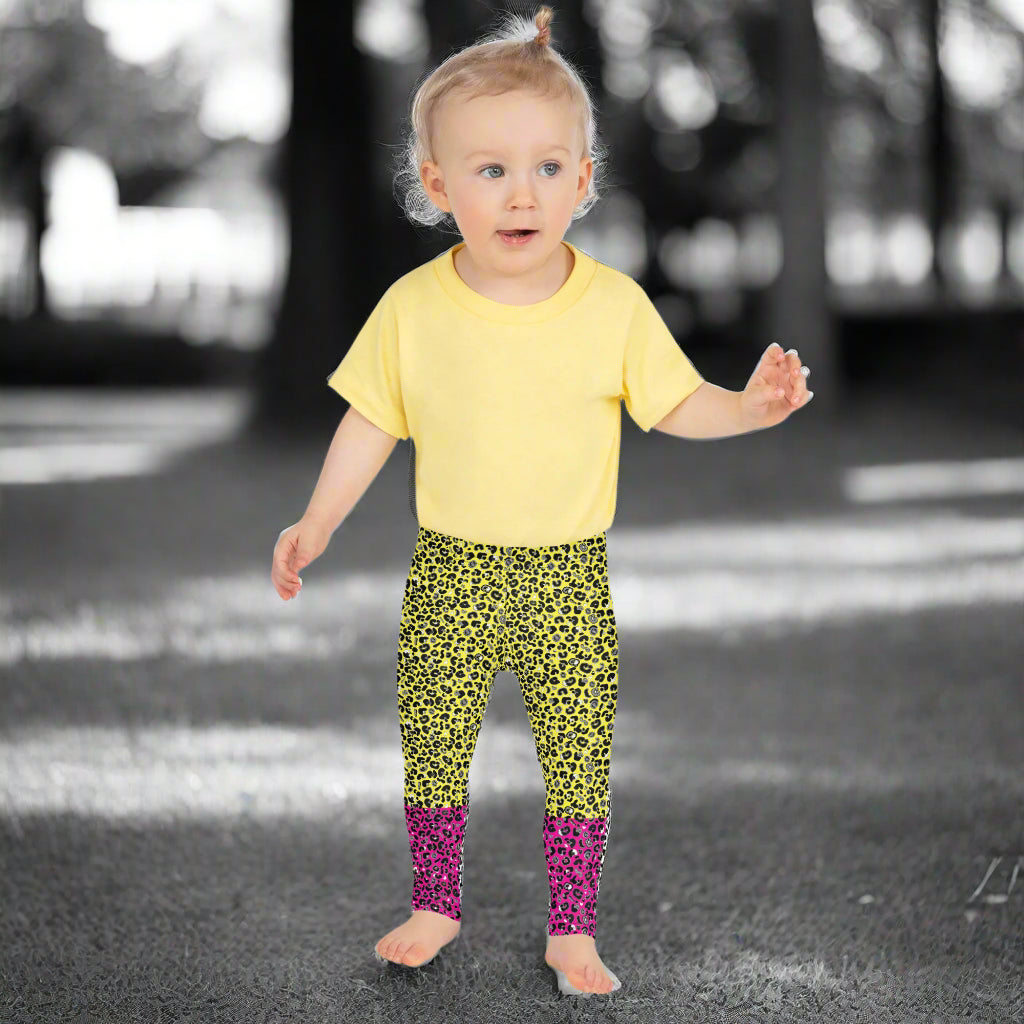A toddler wearing kids size yellow and pink leopard glamour funky festival leggings and a pale yellow t-shirt stands facing forwards. She is standing in front of a black and white park scene scape.  This design features an animal print pattern in black, grey and white, printed at a medium scale. The top 3/4 background is bright yellow with the bottom 1/4 bright pink