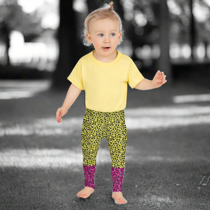 A toddler wearing kids size yellow and pink leopard glamour funky festival leggings and a pale yellow t-shirt stands facing forwards. She is standing in front of a black and white park scene scape.  This design features an animal print pattern in black, grey and white, printed at a medium scale. The top 3/4 background is bright yellow with the bottom 1/4 bright pink