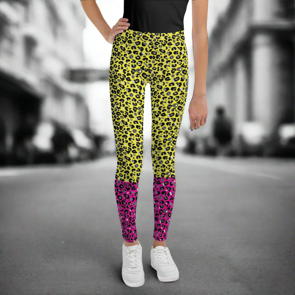 A young person from the waist down wearing youth size yellow and pink leopard glamour funky festival leggings stands facing forwards. They are wearing white trainers and have one hand on their hip. They are standing in front of a black and white city street scape.  This design features an animal print pattern in black, grey and white, printed at a medium scale. The top 3/4 background is bright yellow with the bottom 1/4 bright pink