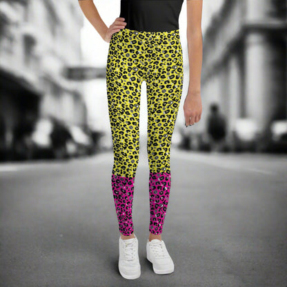A young person from the waist down wearing youth size yellow and pink leopard glamour funky festival leggings stands facing forwards. They are wearing white trainers and have one hand on their hip. They are standing in front of a black and white city street scape.  This design features an animal print pattern in black, grey and white, printed at a medium scale. The top 3/4 background is bright yellow with the bottom 1/4 bright pink