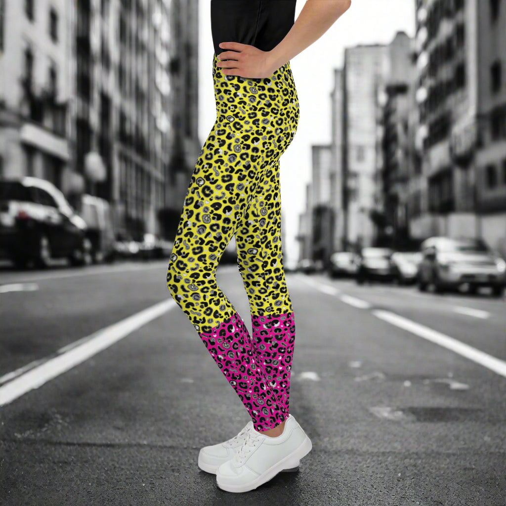 A young person from the waist down wearing youth size yellow and pink leopard glamour funky festival leggings stands facing to the left. They are wearing white trainers and have one hand on their hip. They are standing in front of a black and white city street scape.  This design features an animal print pattern in black, grey and white, printed at a medium scale. The top 3/4 background is bright yellow with the bottom 1/4 bright pink