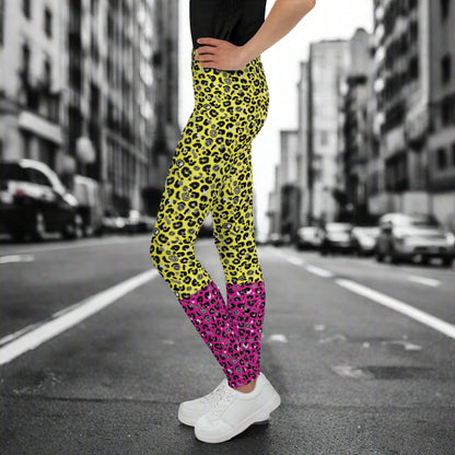A young person from the waist down wearing youth size yellow and pink leopard glamour funky festival leggings stands facing to the left. They are wearing white trainers and have one hand on their hip. They are standing in front of a black and white city street scape.  This design features an animal print pattern in black, grey and white, printed at a medium scale. The top 3/4 background is bright yellow with the bottom 1/4 bright pink