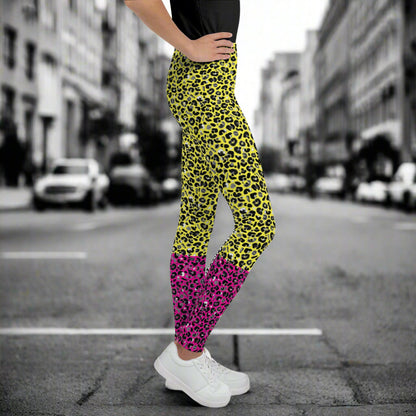 A young person from the waist down wearing youth size yellow and pink leopard glamour funky festival leggings stands facing to the right. They are wearing white trainers and have one hand on their hip. They are standing in front of a black and white city street scape.  This design features an animal print pattern in black, grey and white, printed at a medium scale. The top 3/4 background is bright yellow with the bottom 1/4 bright pink